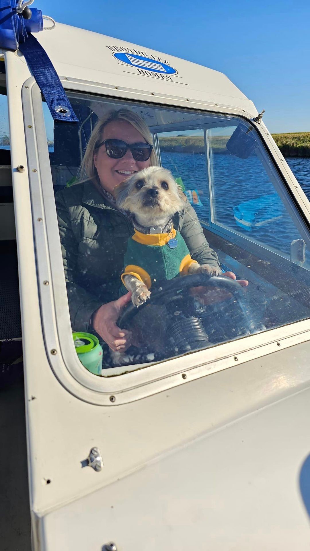 Woman in sunglasses and dog in sweater smiling while steering a boat on a sunny day.